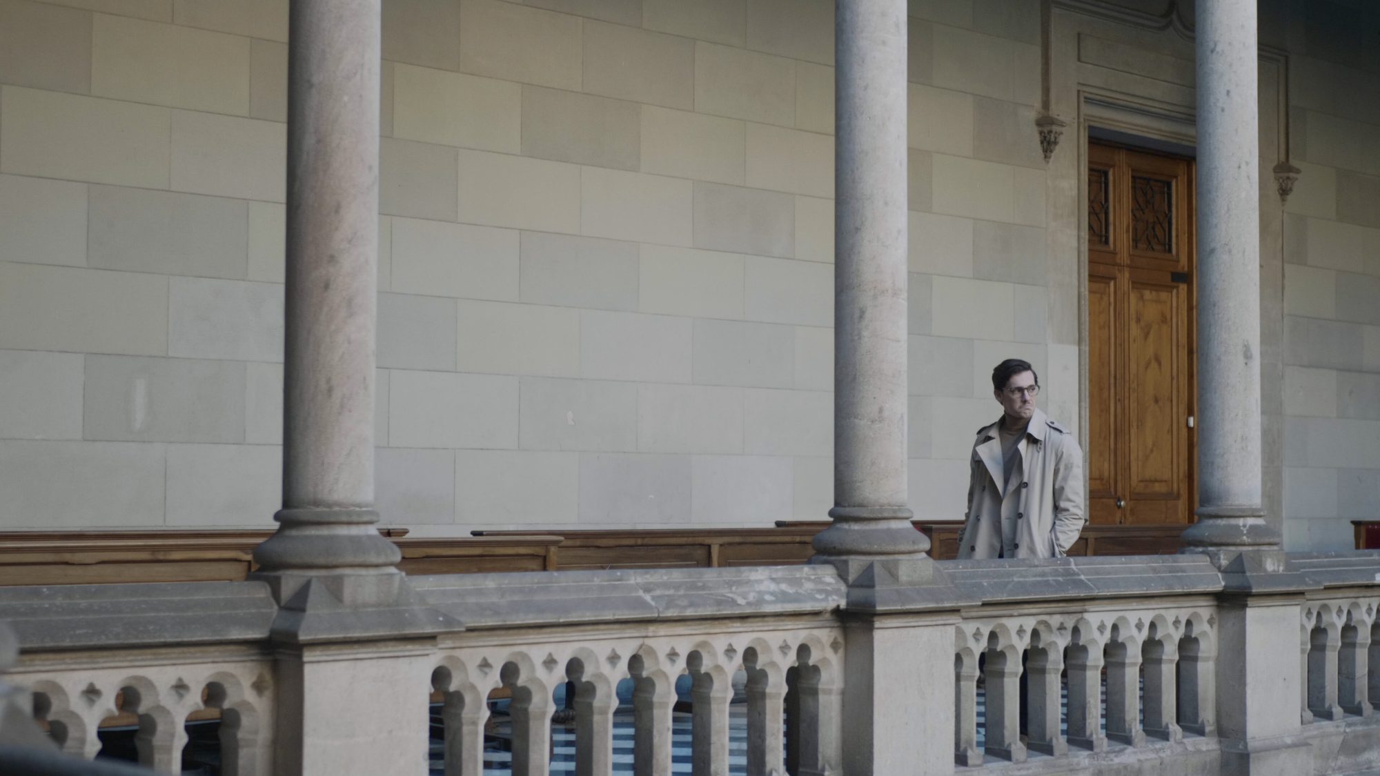 Man in a light coat walking along a classical stone colonnade
