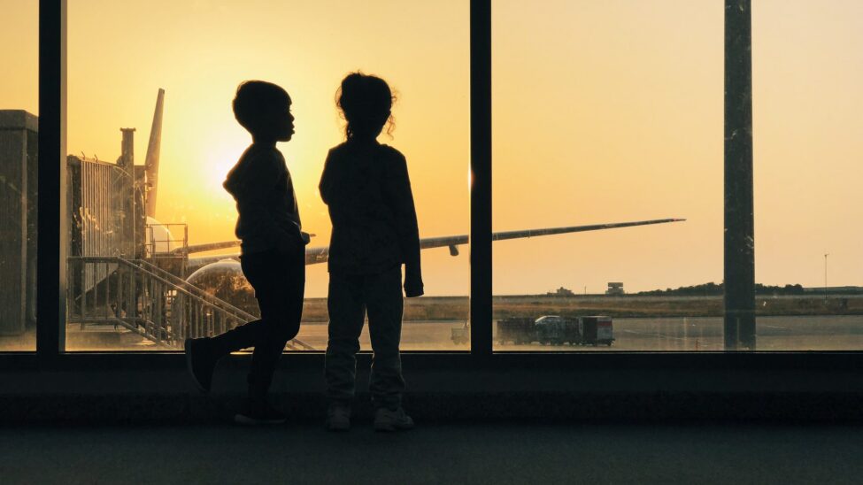 Two children at an airport window at dusk with an aircraft beyond, an emigration moment