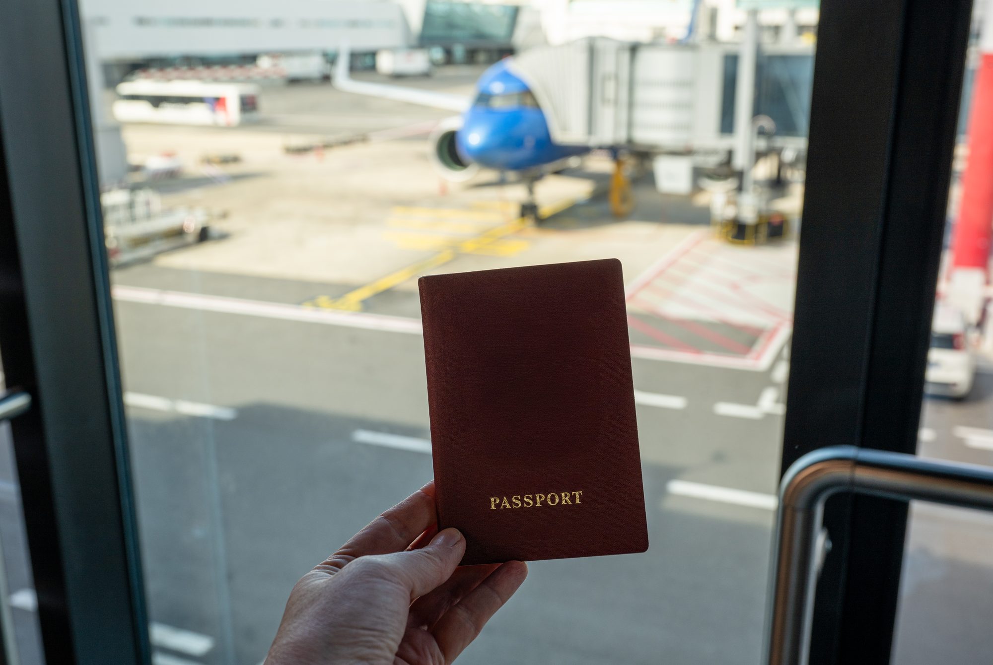 A hand holding a passport at an airport window, with an aircraft at the gate beyond