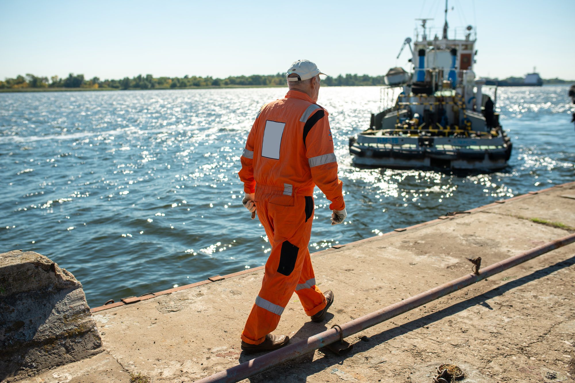 A port worker in orange overalls walking along a quay towards a tug