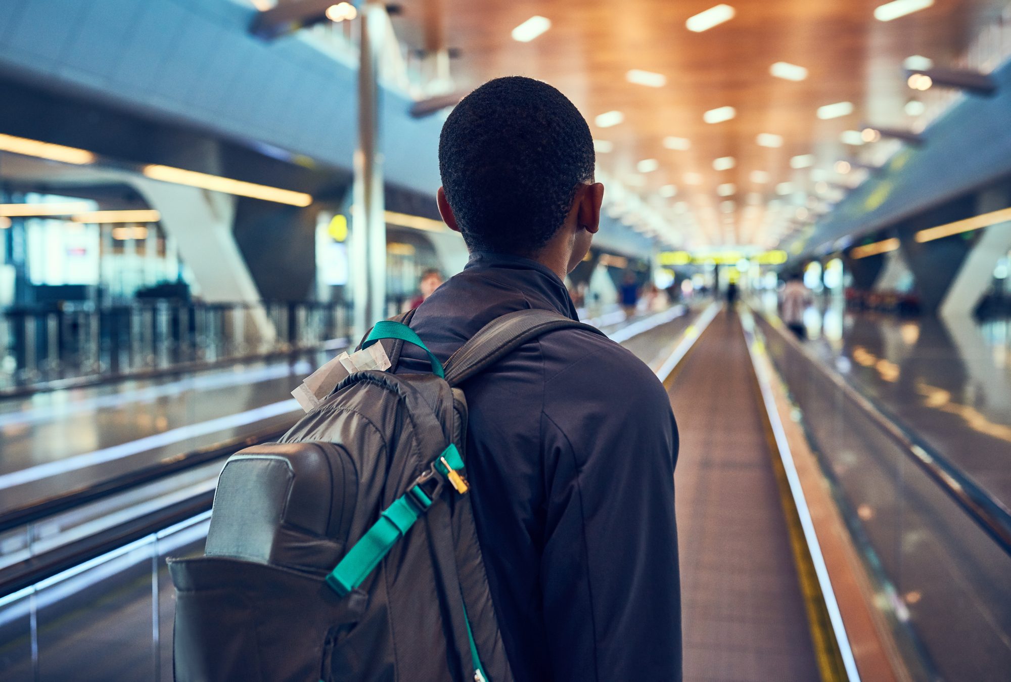A traveller with a backpack on a moving walkway in an airport concourse, seen from behind