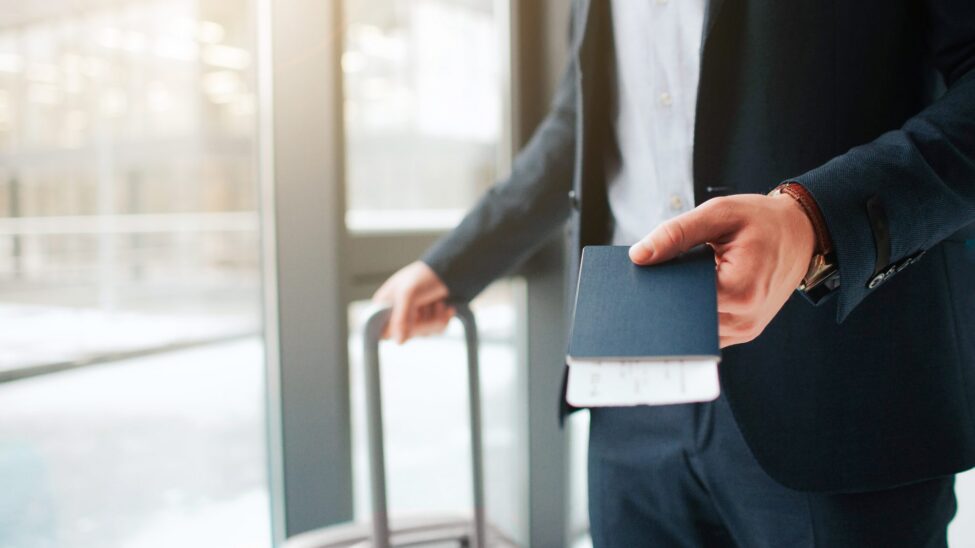 A traveller in a navy suit holding a passport and boarding pass, with a suitcase beside a window